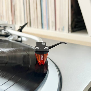 Turntable with a cartridge on a vinyl record, with records in the background.