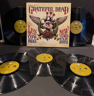Grateful Dead 'Live at the Cow Palace' vinyl record set with the illustrated album cover standing upright behind four black vinyl records on a dark surface.