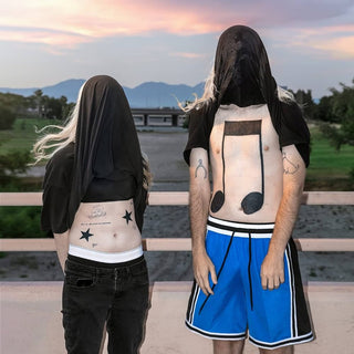 Two people with visible tattoos on their backs standing outdoors with a scenic background.