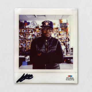 A square instant-film-style photo print featuring a man in a black puffer jacket and a cap with 'Austin Texas' on it, standing in front of shelves filled with memorabilia. The print has a white border with a black autograph at the bottom left and a '2024 Austin Creek Concerts' logo at the bottom right, shown against a light gray textured background.