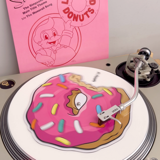 A vinyl record player with a pink record sleeve featuring 'The Simpsons' and 'Lard Lad Donuts' artwork, alongside a pink donut-shaped vinyl skin or decal resting on the turntable.