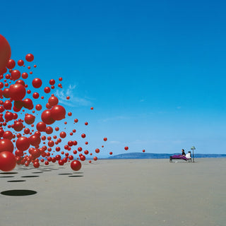 A surreal outdoor scene on a wide sandy beach under a bright blue sky, with many red spheres floating or standing in the left side and a small bench with two people on the right.