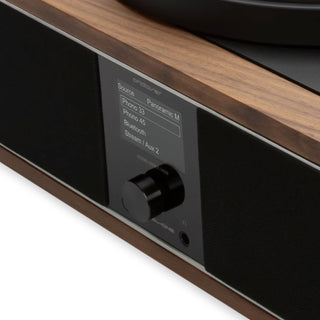 Close-up of a speaker system control panel with a wood-grain top, black speaker grilles, and a central display screen showing Bluetooth and source options.