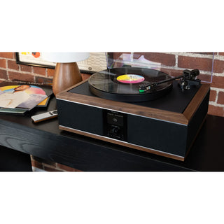 A black and wood turntable with a vinyl record on it, placed on a dark table in a room setting with a brick wall, lamp, and records in the background.