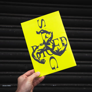 A hand holds a bright yellow rectangular card or poster featuring a black graphic design with stylized text and a snake illustration, set against a dark corrugated metal background.