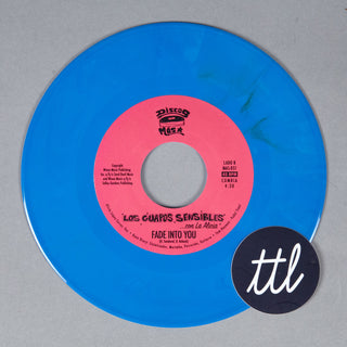 A translucent blue vinyl record with a pink center label reading 'Los Guapos Sensibles - Son La Alameda' and 'Fade Into You,' photographed on a light gray background. A black circular sticker with white 'ttl' script is placed in front of the record.