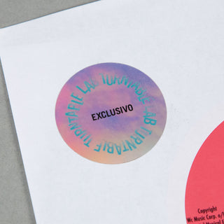 A close-up of a round holographic sticker on a white sheet of paper, featuring a purple, pink, and blue iridescent gradient with the text 'EXCLUSIVO' in black and mirrored Spanish text around the edge.
