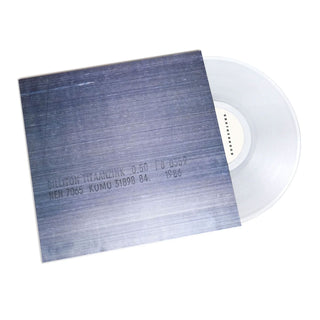 A translucent clear vinyl record partially pulled from a square album sleeve with a blue-gray brushed-metal style cover, photographed on a white background.
